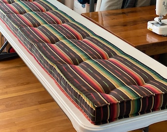 Puede incluir: Un cojín de banco largo y rectangular con una tela a rayas marrón, verde, roja y amarilla. El cojín tiene múltiples botones y está sentado en una mesa blanca.