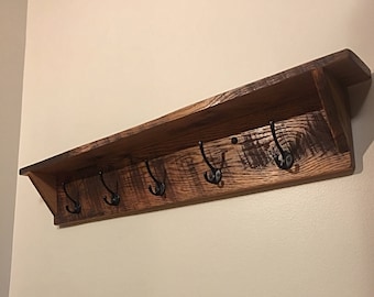 Puede incluir: Una estantería de madera rústica con cinco ganchos de metal negro. La estantería tiene un acabado desgastado y está montada en una pared.