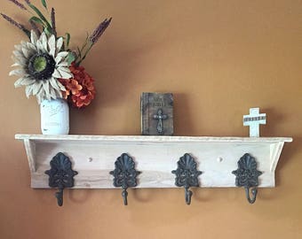 Puede incluir: Una estantería de madera blanca con cuatro ganchos de metal negro. La estantería tiene un acabado de madera clara y está montada en una pared. Hay un pequeño libro titulado "Holy Bible" y una pequeña cruz en la estantería.
