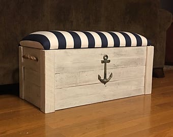 Puede incluir: Cofre de almacenamiento de madera blanca con un cojín superior a rayas azules y blancas y un acento de ancla de metal.