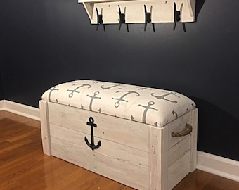 Puede incluir: Banco de almacenamiento de madera blanca con un cojín de patrón de ancla gris y blanco. El banco tiene un asa de cuerda y un ancla de metal negro en la parte delantera.