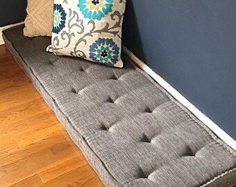 Puede incluir: Un cojín de banco gris con mechones y ribete blanco, colocado sobre un suelo de madera, con dos cojines decorativos. Uno es beige con un patrón texturizado, el otro tiene diseños florales azules y verdes.