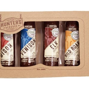 Op de afbeelding: Een kartonnen doos met een transparant venster dat vier verschillende soorten gerookte zomerworst laat zien: bizon, hert, eland en wild zwijn. De worst is verpakt in doorzichtig plastic met een bruin etiket waarop "Hunter's Reserve" staat.