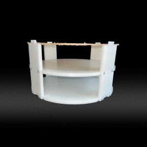 May include: A white, two-tiered, circular display stand. The stand has a simple design with two shelves and vertical supports. The edges are slightly textured, and small rivets are visible. The top has a thin, light brown strip.