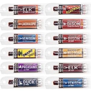 Op de afbeelding: Twaalf individueel verpakte vleesstokjes met verschillende wildsmaken, waaronder eland, jackalope, wild zwijn, swamp stick, fazant, eend, bizon, roadkill en wild zwijn. Elke verpakking heeft een kleurrijk ontwerp met de naam van het vleesstokje en een kleine illustratie.