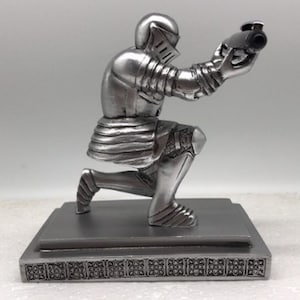 Op de afbeelding: Een zilveren ridderfiguur knielt op een voetstuk met een decoratieve rand. De ridder houdt een zilveren pen in zijn rechterhand.