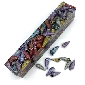 May include: A resin pen blank with iridescent seashells embedded in clear resin. The blank is rectangular and has a smooth, polished surface. There are also several loose seashells scattered around the blank.