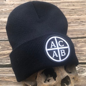 May include: A black beanie with a white circle patch that says "ACAB" on it. The beanie is sitting on a skull.