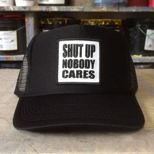 May include: Black trucker hat with a white square patch that says "SHUT UP NOBODY CARES" in black letters.