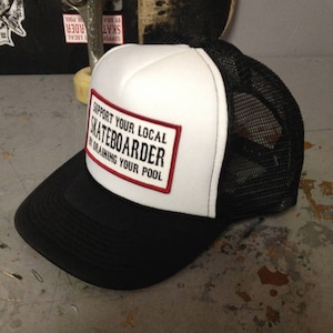 Puede incluir: Gorra de camionero blanca y negra con un parche rojo y blanco que dice "Support Your Local Skateboarder By Draining Your Pool".