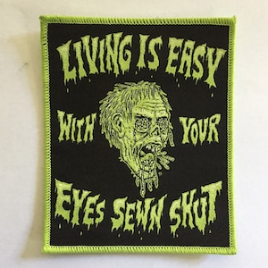 May include: Black square patch with a neon green border and text that reads "LIVING IS EASY WITH YOUR EYES SEWN SHUT." A zombie-like illustration is in the center. The patch is likely for clothing or accessories.