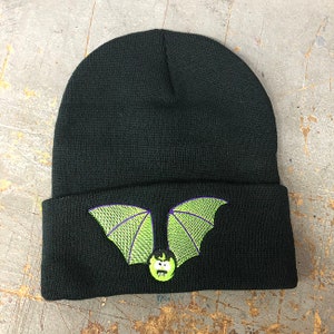 May include: Black knit beanie with a green and purple embroidered bat design. The bat has a cartoon face with a green complexion and a wide, toothy grin.