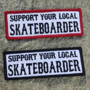 May include: Two embroidered patches with the text "Support Your Local Skateboarder". The top patch has a red border and white background with black text. The bottom patch has a black border and white background with black text.