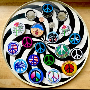 May include: A collection of colorful peace symbol buttons and pins, some with text like "Make Love Not War" and "50 Years Old Still Going Strong." The buttons are arranged on a black and white spiral background.