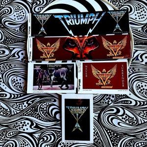 Könnte beinhalten: Eine Sammlung von Triumph-Bandaufklebern und Mini-Postern. Die Aufkleber zeigen das Bandlogo und die Wörter "TRIUMPH" und "SURRENDER". Die Mini-Poster zeigen Bilder der Band bei Live-Auftritten. Der Hintergrund ist eine schwarz-weiß gemusterte Oberfläche.