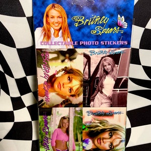 May include: Collectible photo stickers featuring images of Britney Spears. The packaging is blue with the singer's name in gold script and a butterfly graphic. The stickers showcase various photos of Spears.