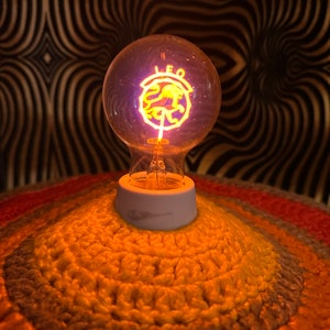 May include: A glowing light bulb with the word "LEO" and a lion symbol. The bulb sits on a white base atop a crocheted, multi-colored surface. The background features a black and gold wavy pattern.