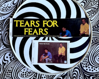 Può includere: Due adesivi vintage e un mini poster con la band Tears for Fears. Gli adesivi e il poster sono su un piatto a strisce bianche e nere, su uno sfondo a motivi bianchi e neri. Il nome della band è in testo giallo.