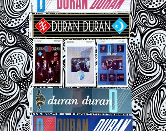 Può includere: Una collezione di adesivi e poster dei Duran Duran. Gli adesivi presentano il nome della band in vari caratteri e colori, tra cui rosso, blu e oro. Alcuni poster mostrano foto della band. Lo sfondo è un motivo a spirale in bianco e nero.