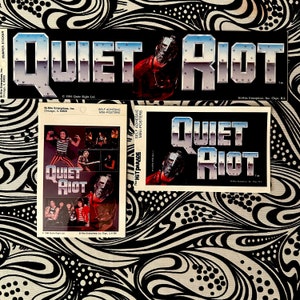 May include: A collection of vintage "Quiet Riot" band merchandise, including a bumper sticker and mini-posters. The items feature the band's logo in a metallic font and an image of a band member in a mask. The background is black and white.