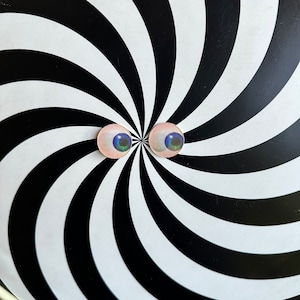 May include: Two round eye-shaped stickers with blue and green irises and pink skin tones are centered on a black and white spiral background. The spiral pattern creates an optical illusion.