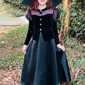 May include: A black witch costume with a long skirt and a velvet bodice with decorative buttons. The bodice has sheer, puffed sleeves and a lace panel. A wide-brimmed black hat completes the outfit, suitable for Halloween or fancy dress events.