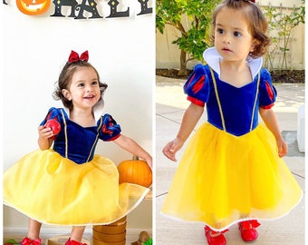 May include: A young child dressed as Snow White for Halloween. The costume features a blue velvet bodice with a yellow tulle skirt, red sleeves, and a yellow sash. The child is wearing a red bow in their hair and holding a red apple.