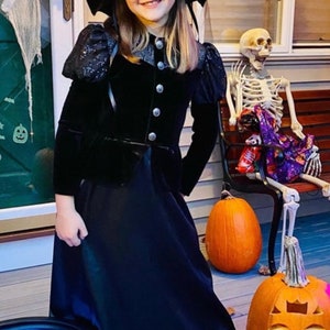 May include: A person in a black witch costume, including a hat and dress, smiles at the camera. The Halloween scene includes carved pumpkins, a skeleton, and a cauldron. The costume has silver buttons.