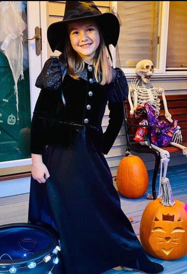 May include: A person in a black witch costume, including a hat and dress, smiles at the camera. The Halloween scene includes carved pumpkins, a skeleton, and a cauldron. The costume has silver buttons.