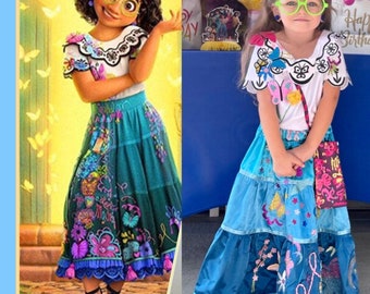 May include: A young girl dressed as the character Mirabel from the Disney film Encanto. She is wearing a blue and green skirt with butterflies and flowers embroidered on it. She is also wearing a white shirt with a butterfly collar and green glasses.
