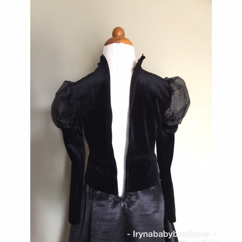 May include: A black velvet jacket with sheer, dotted sleeves and a high collar. The jacket is displayed on a mannequin, showcasing its full-length zip and tailored design. The bottom portion of the jacket is attached to a black satin skirt.