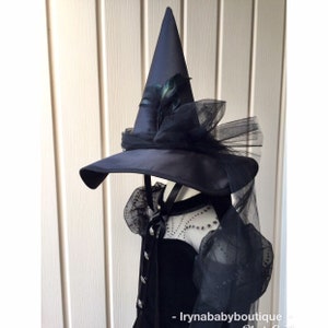 May include: A navy blue witch's hat with a wide brim, decorated with black tulle and feathers. The hat is on a black dress with sheer sleeves and decorative buttons, suitable for a gothic or Halloween costume.