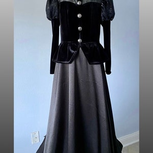 May include: A black velvet and satin gown on a mannequin. The bodice has a high neckline with sheer detailing, sequined sleeves, and decorative buttons. The skirt is a flowing, floor-length design.