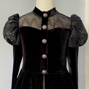 May include: Black velvet jacket with a high collar and sequined puff sleeves. The jacket features a sheer panel across the shoulders and decorative silver buttons down the front. The bottom of the jacket has a peplum design.