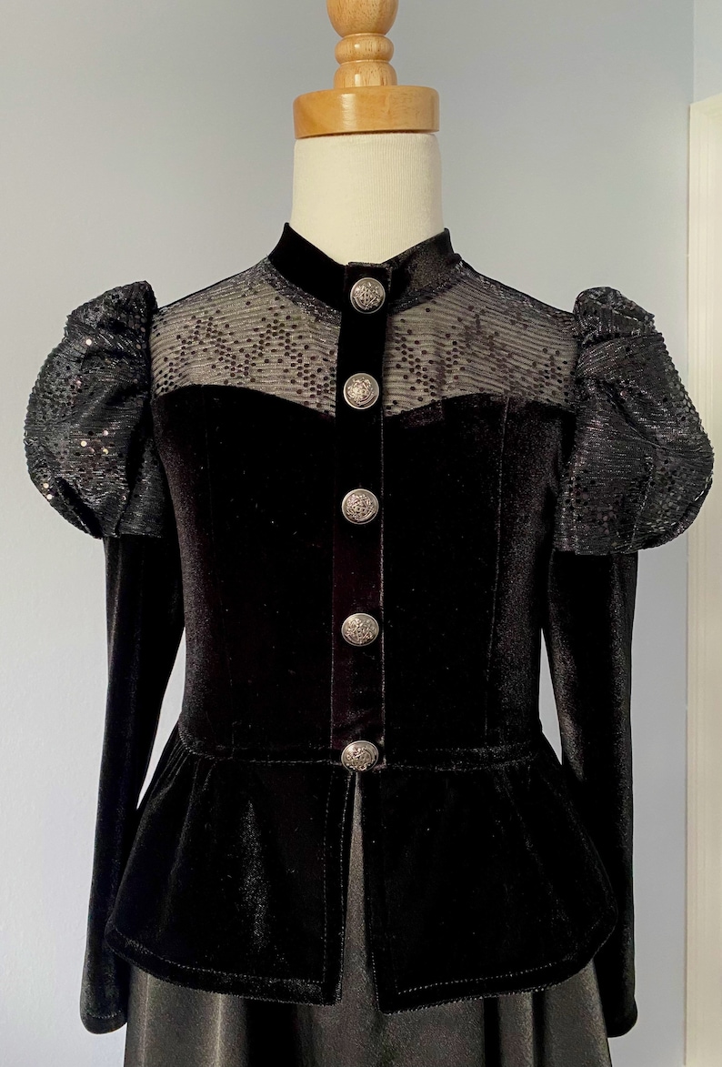 May include: Black velvet jacket with a high collar and sequined puff sleeves. The jacket features a sheer panel across the shoulders and decorative silver buttons down the front. The bottom of the jacket has a peplum design.