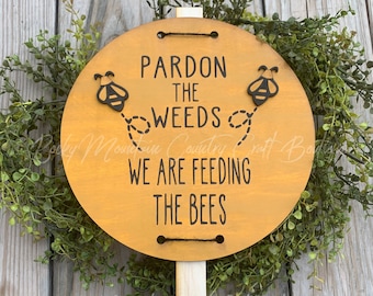 May include: A wooden sign with a yellow background and black text that reads "Pardon the weeds, we are feeding the bees". Two black and yellow bee cutouts are attached to the sign.