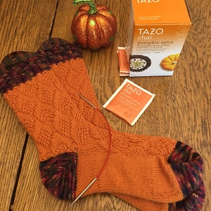 May include: A pair of orange knitted socks with a multi-coloured heel and toe. The socks are lying on a wooden surface with a small pumpkin and a box of Tazo pumpkin spice chai tea bags.