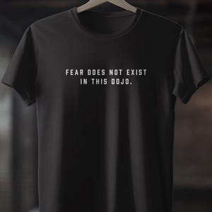 May include: Black t-shirt with the white text "FEAR DOES NOT EXIST IN THIS DOJO." The shirt is hanging on a wooden hanger. The t-shirt is made of a soft material.