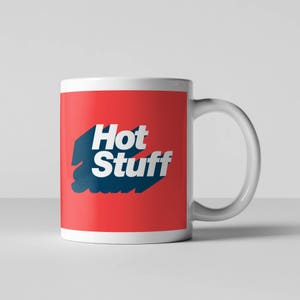 Op de afbeelding: Witte keramische mok met een rode achtergrond en de tekst "Hot Stuff" in blauwe letters.