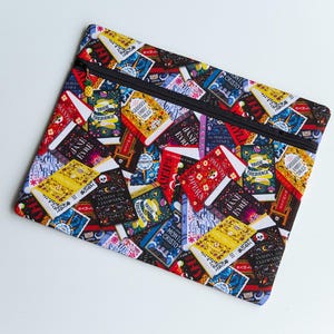 May include: A rectangular fabric zip-up pouch featuring a colourful all-over print of various classic book covers, including titles like Jane Eyre, Monte Cristo, and Pride and Prejudice. The pouch has a black zip closure with a small pineapple charm.