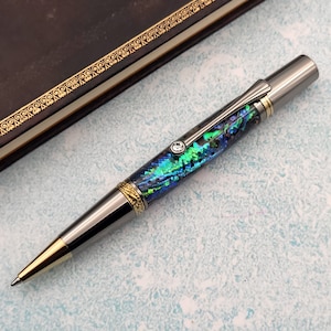Puede incluir: Un bolígrafo negro y dorado con un cuerpo de resina iridiscente azul y verde. El bolígrafo tiene un acento en forma de diamante cerca de la parte superior del cuerpo.