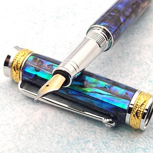 Puede incluir: Una pluma estilográfica azul y verde con detalles plateados y dorados. La pluma tiene una punta fina y un capuchon que se atornilla.