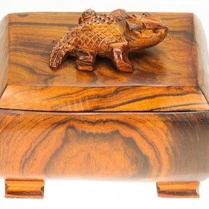 Peut inclure: Une boîte en bois avec un lézard sculpté sur le couvercle. La boîte est en bois foncé avec une teinte brun rougeâtre et a une forme carrée aux bords arrondis. La boîte a quatre petits pieds.