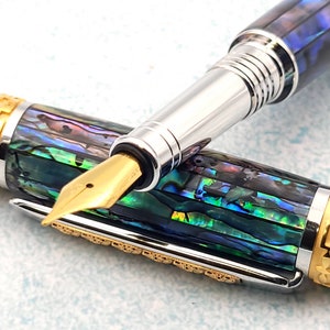 Op de afbeelding: Een gouden en zilveren vulpen met een blauw en groen abalone-schelpontwerp. De pen heeft een vergulde penpunt en een draaimechanisme.