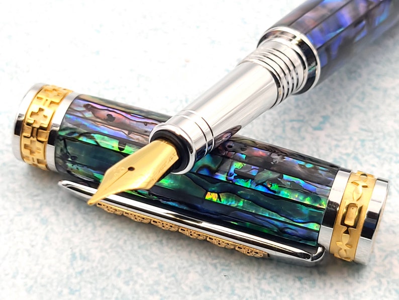 Op de afbeelding: Een gouden en zilveren vulpen met een blauw en groen abalone-schelpontwerp. De pen heeft een vergulde penpunt en een draaimechanisme.