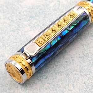 Op de afbeelding: Een close-up van een blauwe en groene abalone-schelp pen met gouden accenten. De pen heeft een zilveren clip en een gouden band met een decoratief patroon.