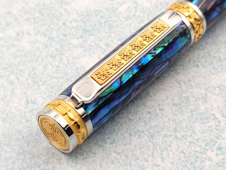 Op de afbeelding: Een close-up van een blauwe en groene abalone-schelp pen met gouden accenten. De pen heeft een zilveren clip en een gouden band met een decoratief patroon.