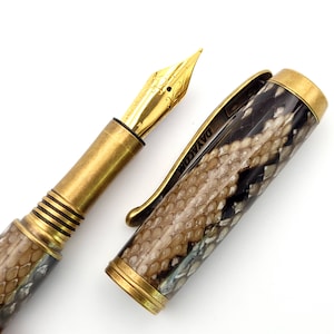 Può includere: Una penna stilografica di lusso con pennino in oro e dettagli in ottone. Il fusto e il cappuccio della penna presentano un motivo a pelle di serpente nei toni del marrone, nero e crema. La clip della penna riporta la scritta "DAYA.COM".