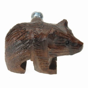 May include: A wooden bear bottle stopper with a brown finish. The bear is carved in a standing position with its head turned to the side. The stopper has a metal screw top.
