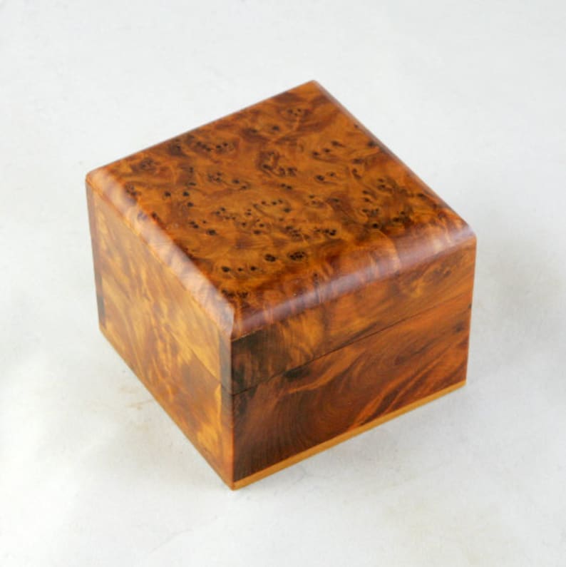 May include: A small, square wooden box with a natural wood grain finish. The box has a hinged lid and is made of a light brown wood.